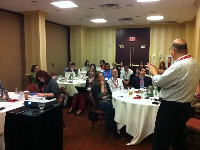 There are many people sitting around a table in their chairs. Some of them are placing laptops in...