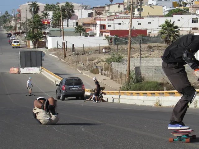 In this image there is a road. There are people skating on the road. There are vehicles parked. In...