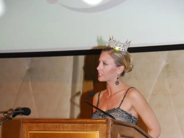 In the image we can see a woman wearing clothes, earrings and a crown. There is a podium and this...