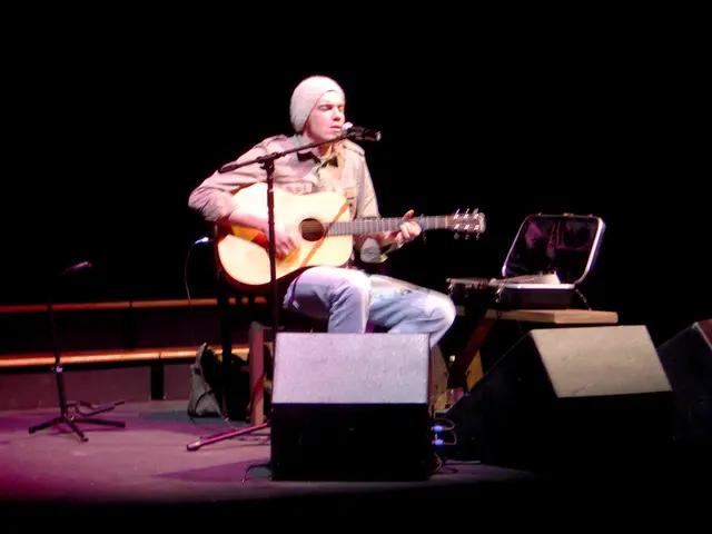 In this image I can a person sitting on the stage and this person is playing guitar and singing.