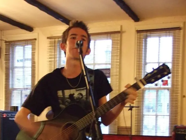 Here is a man wearing the black T-shirt,trouser and standing. He is playing guitar and singing...