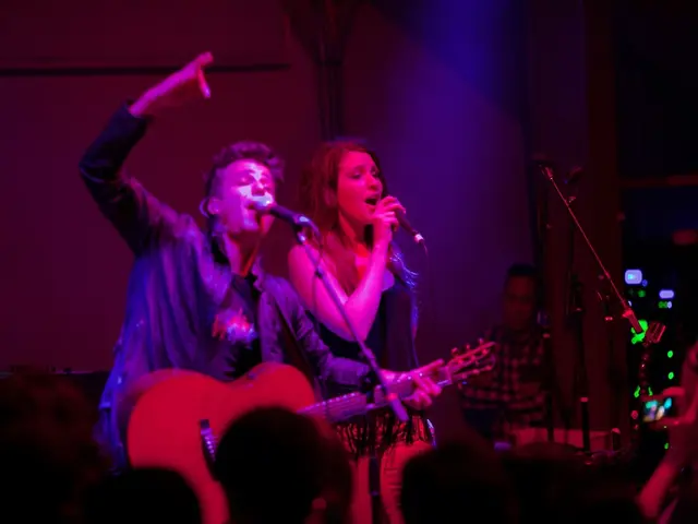 On the stage there are three people. To the left side there is a man with black jacket standing and...