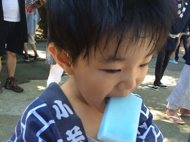 In this image I can see a kid is licking the ice-cream, this kid wore blue color cloth. Behind this...