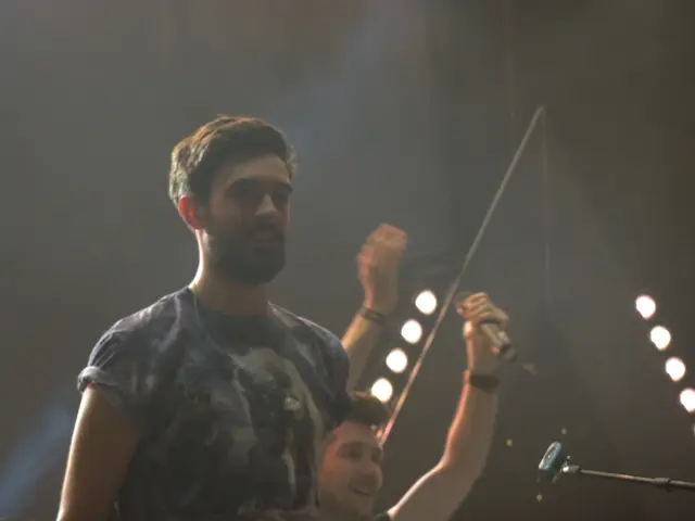 There is a man in t-shirt, laughing and standing. In the background, there is another man laughing,...