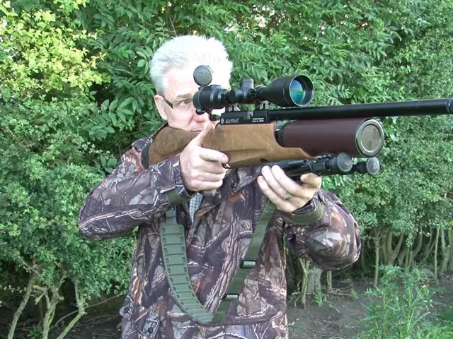 In the image there is a man standing and holding a gun in his hand. Behind him there are trees.