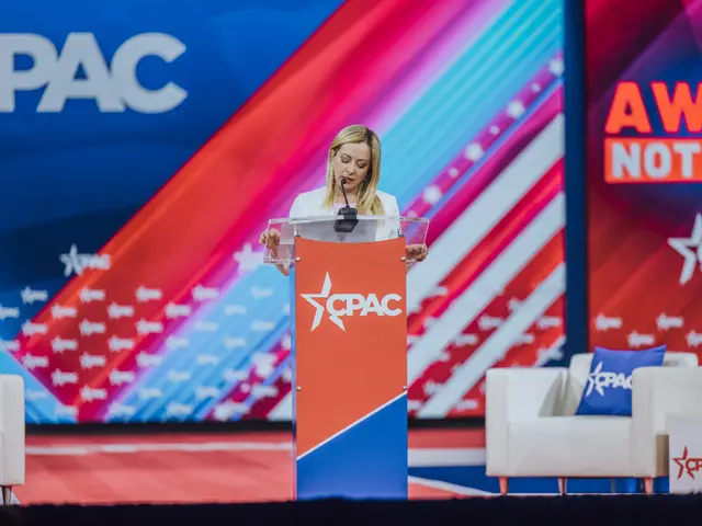 The image shows a woman standing at a podium in front of a screen, with a microphone in her hand....