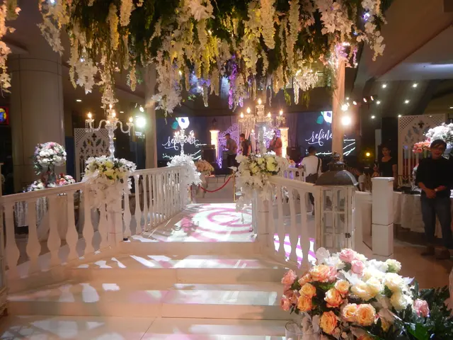 The image shows a wedding ceremony taking place in a large room with white flowers and chandeliers...