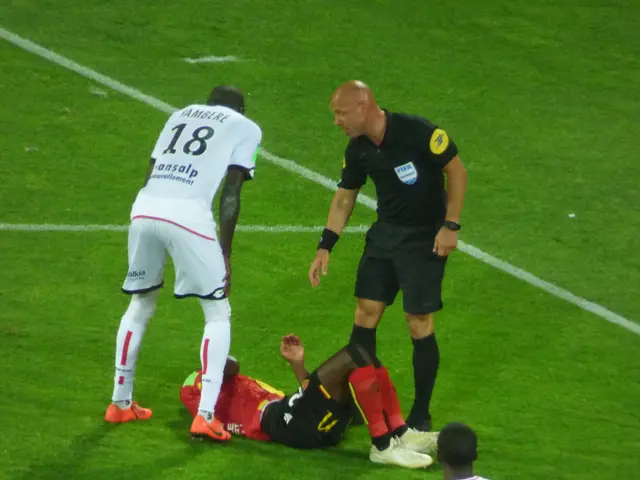 The image shows a soccer player laying on the ground next to a referee, surrounded by three other...