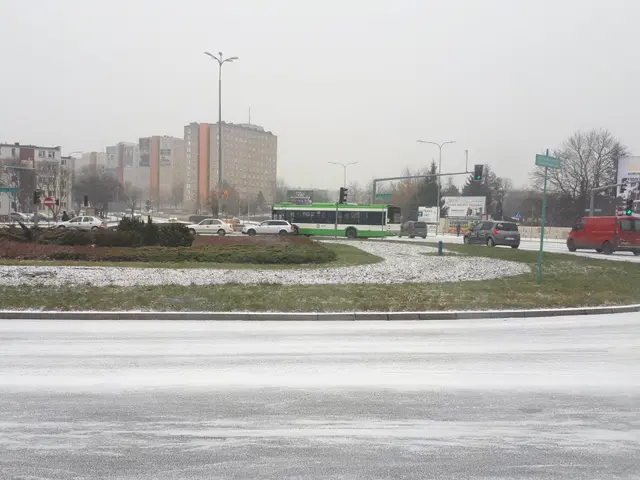The image shows a city street with cars and buses driving down it, surrounded by snow on the...