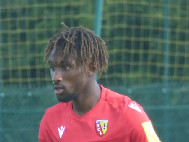The image shows a man with dreadlocks standing on a soccer field, wearing a red t-shirt and shorts....