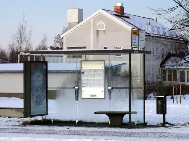The image shows a bus stop in the middle of a snowy street, surrounded by buildings, trees, plants,...