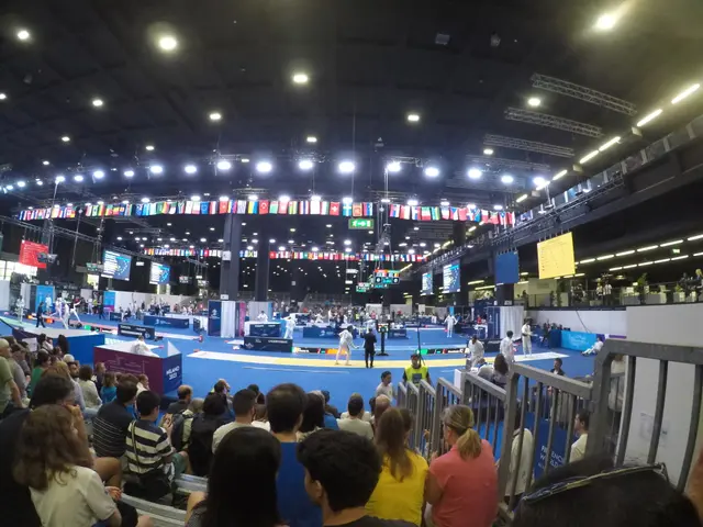 The image shows a large crowd of people sitting in front of a stage, with a railing on the right...