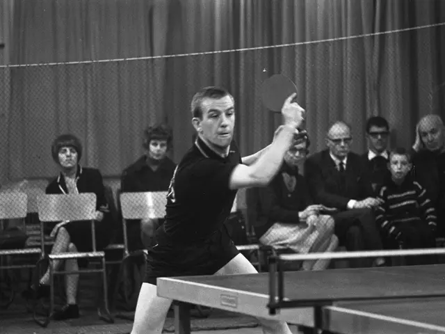 The image shows a man playing ping pong in front of a crowd of people, some of whom are sitting on...