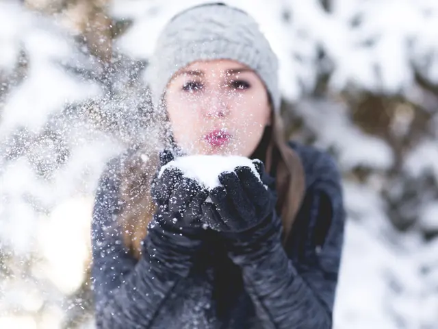 The image shows a woman wearing a cap and gloves, blowing snow in the air. The background is...
