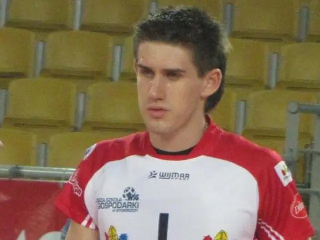 The image shows a man in a red and white uniform standing on a court, surrounded by a few people...