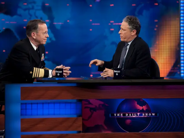 The image shows two men sitting at a table in front of a TV screen, both wearing black blazers,...
