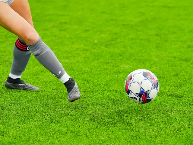 The image shows a woman kicking a soccer ball on top of a lush green field. She is wearing sports...