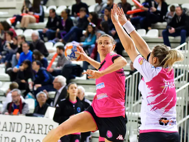 The image shows two women playing a game of volleyball on a court, with a group of people sitting...