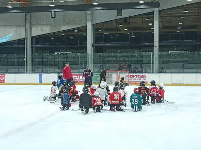 The image shows a group of children sitting on top of an ice rink, wearing helmets and holding...
