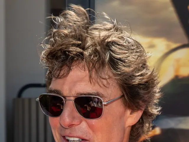 The image shows a man in a suit and tie standing on a red carpet, wearing goggles and smiling. In...