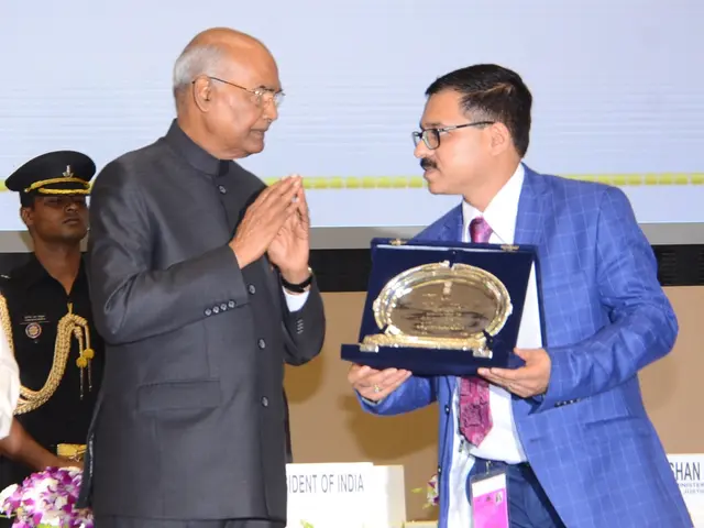 The image shows two men standing side by side on a stage. The man on the right is wearing a blue...