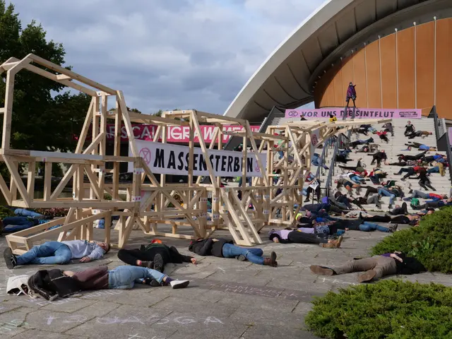 The image shows a group of people laying on the ground in front of a building, surrounded by plants...