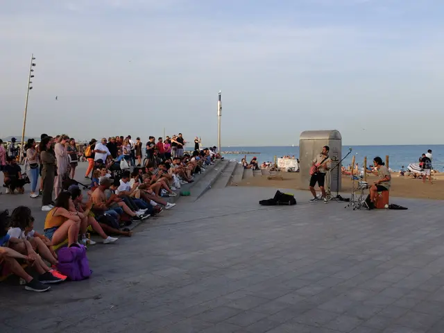 The image shows a group of people sitting on top of a sidewalk next to the ocean. There are men,...