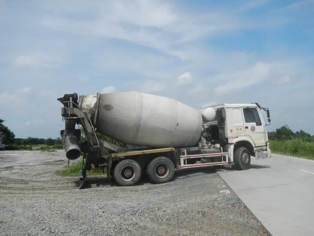 The image shows a cement mixer truck parked on the side of the road, surrounded by stones, grass,...
