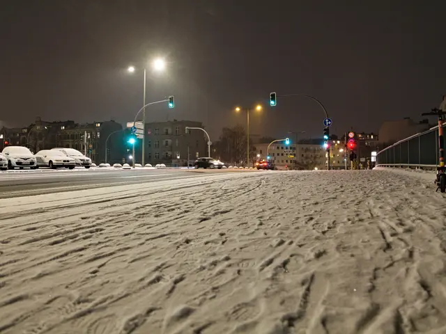 The image shows a snowy street at night with cars parked on the side of it. We can see snow on the...