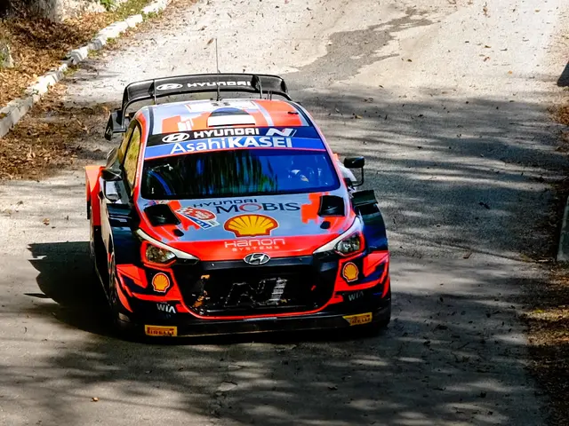 The image shows a rally car driving down a winding road surrounded by trees and plants. The car is...