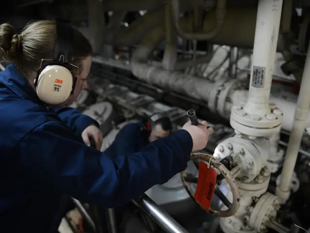 The image shows two women in blue uniforms working on a machine, with one of them holding an object...