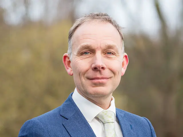 The image shows a man in a blue suit and tie standing in front of a blurred background, smiling. He...