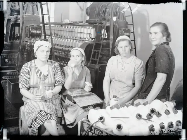The image shows a group of four women sitting around a table in a factory. They are all smiling and...