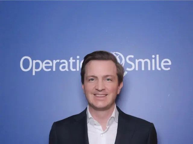 The image shows a man in a suit standing in front of a blue wall with the words "Operationsmile"...