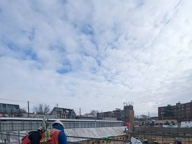 The image shows two construction workers wearing jackets, helmets, and gloves, standing on top of a...
