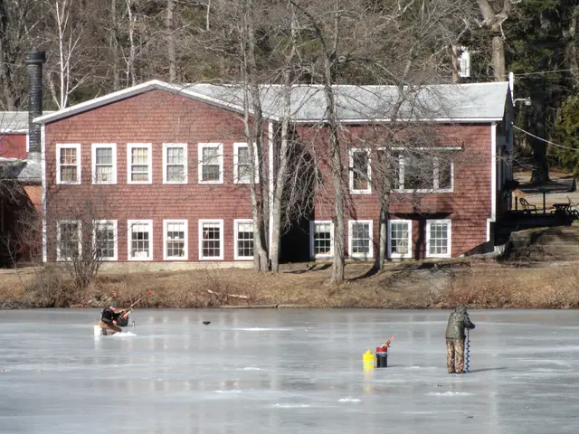 The image shows two people ice fishing on a frozen lake in front of a red brick building. The lake...