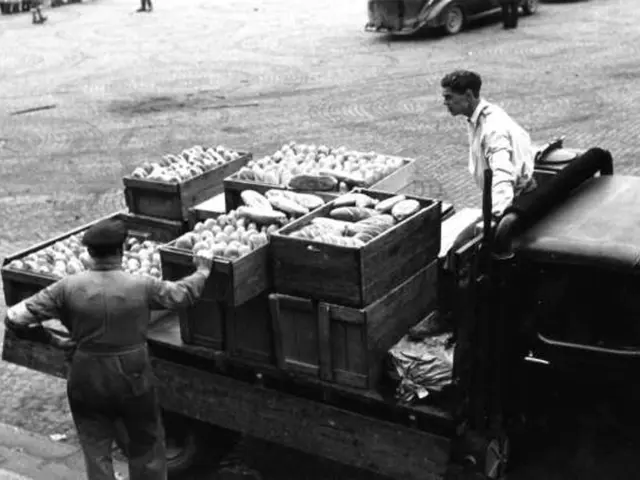 The image shows a black and white scene of two men loading crates of vegetables into the back of a...