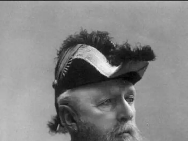 The image shows an old black and white photo of a man with a beard wearing a cap and a medal around...
