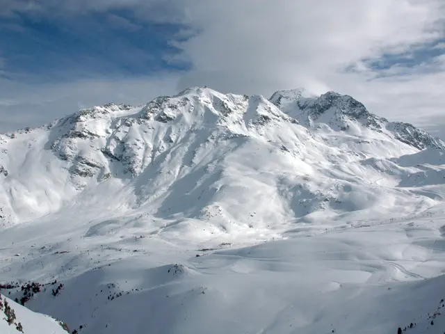 The image shows a snow covered mountain with a few people skiing down it, surrounded by a sky...