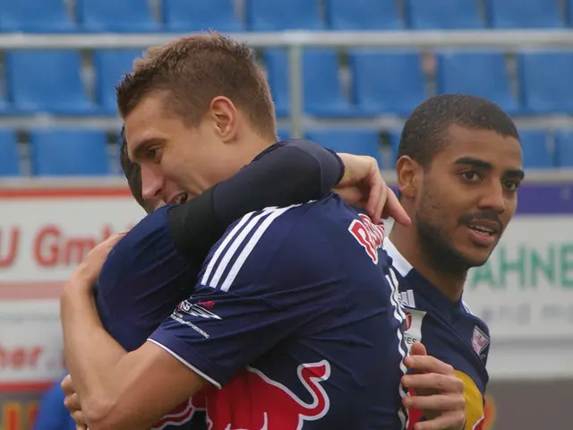 The image shows two soccer players embracing each other on a field, with a person's hand visible on...