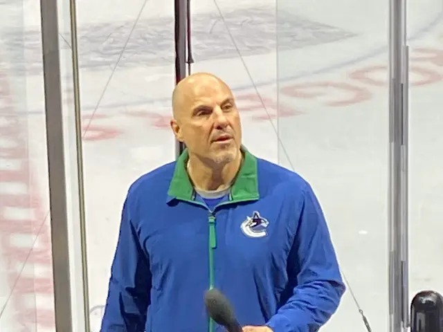 The image shows a man in a blue jacket standing in front of a hockey rink, holding a microphone in...