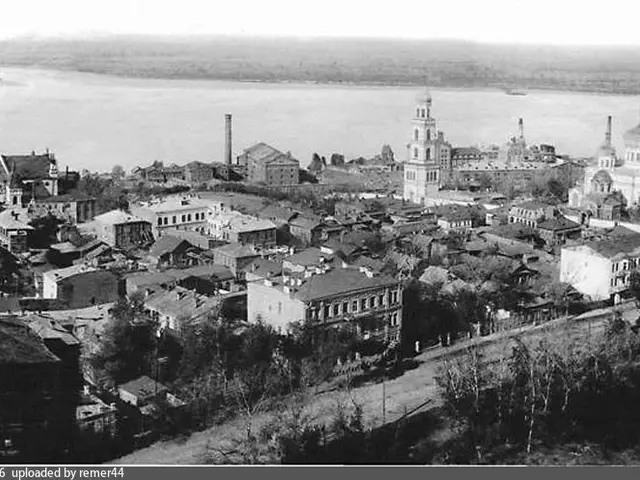 The image shows a black and white photo of a city with many buildings, trees, and a body of water...