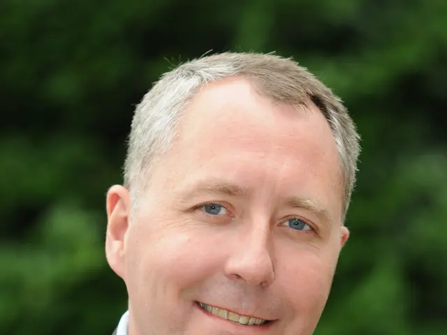 The image shows a man in a suit and tie smiling at the camera with a blurred background. He appears...