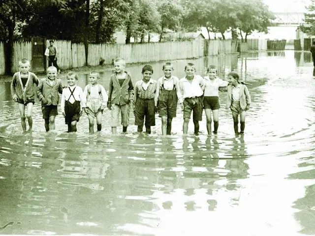 The image shows a group of children walking through a flooded street, with a person standing on the...