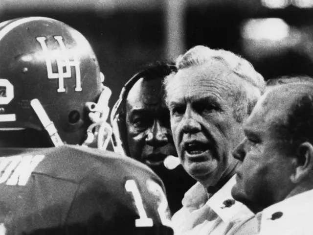The image shows a black and white photo of a football coach talking to his team, with a person...