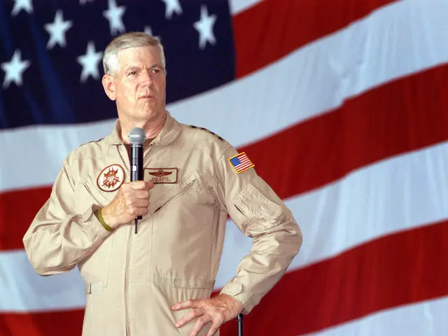 The image shows a man in a military uniform standing in front of an American flag, holding a...