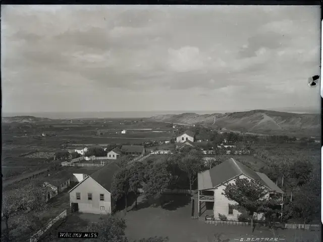 The image shows a black and white photo of a small town with houses, trees, and mountains in the...