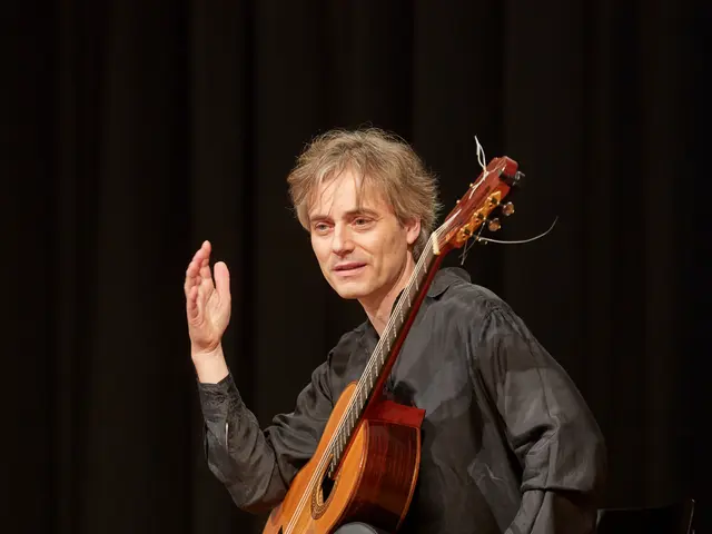 The image shows a man sitting on a chair playing a guitar in front of a black curtain. He is...