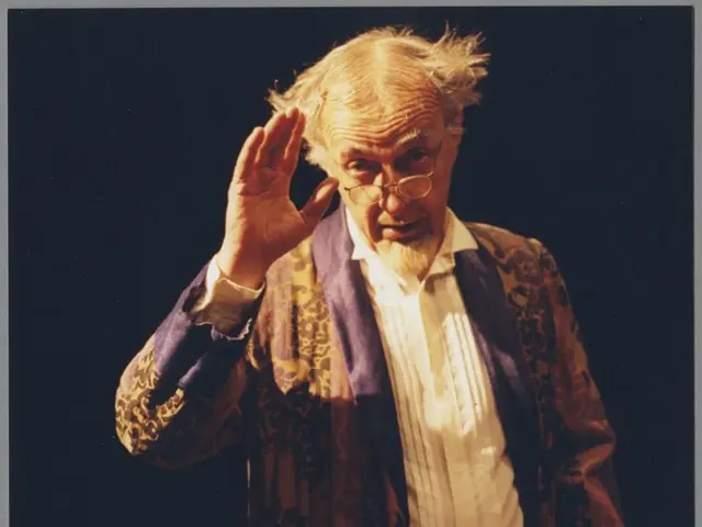 The image shows a man in a robe and glasses standing on a stage, with a dark background. He appears...