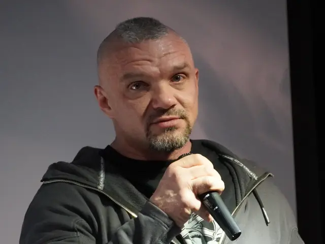 The image shows a man in a black hoodie holding a microphone, standing in front of a wall. He...